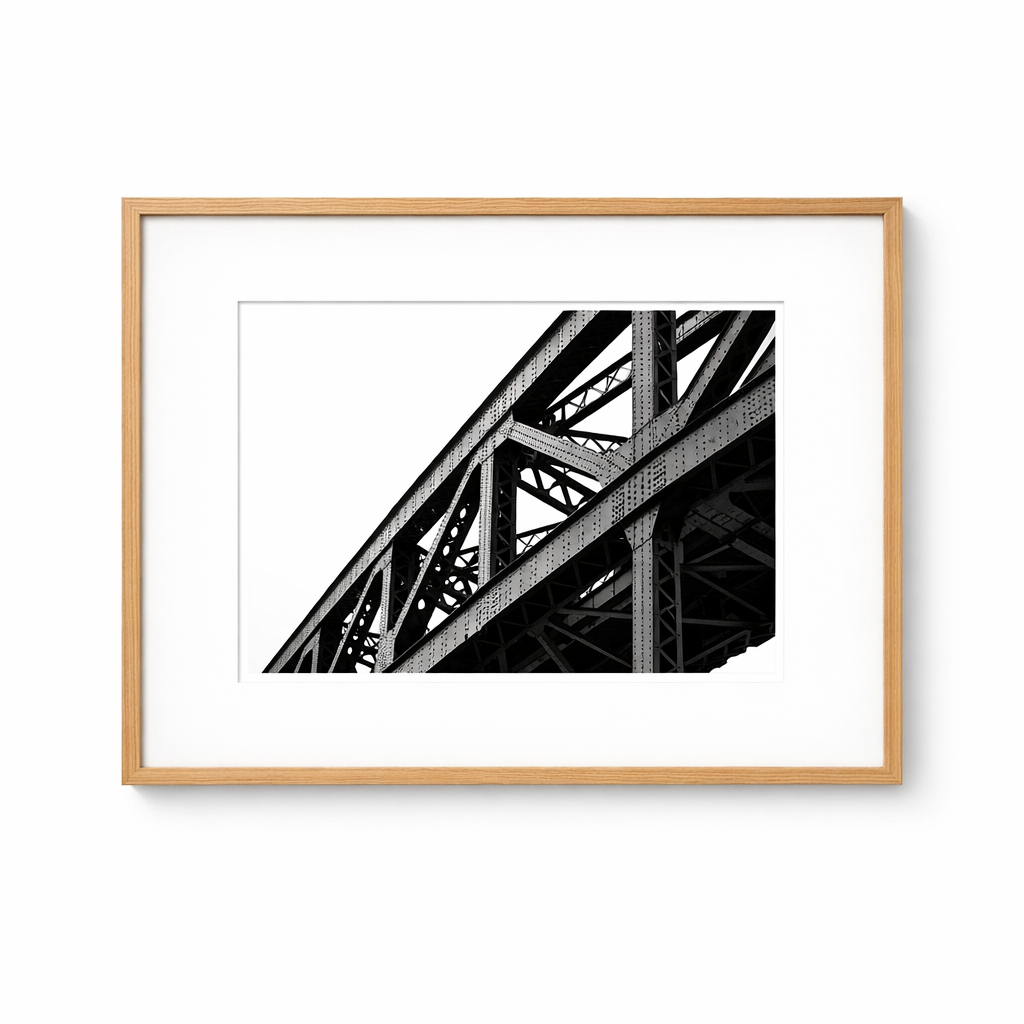 A large, black-and-white architectural print of steel bridge trusses framed in a thin light oak frame, resting against a pure white wall.