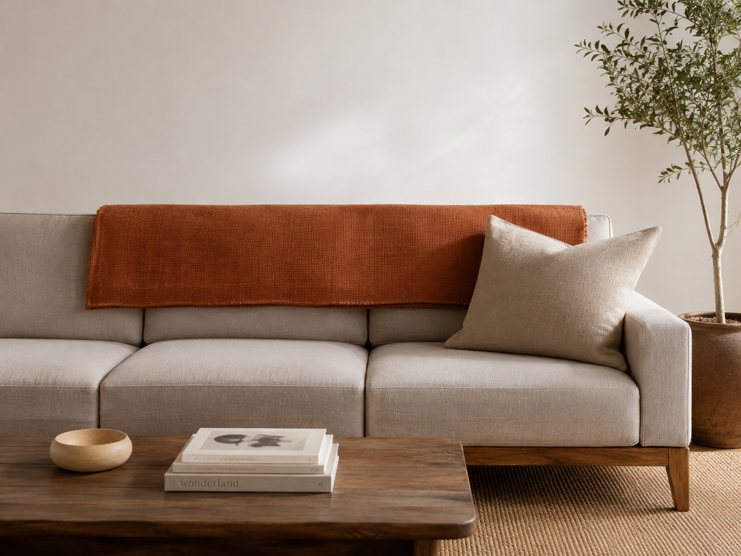 TextA flat-weave terracotta throw folded neatly in a wide horizontal rectangle along the back of a beige sofa, ends tucked under the cushions, editorial interior styling